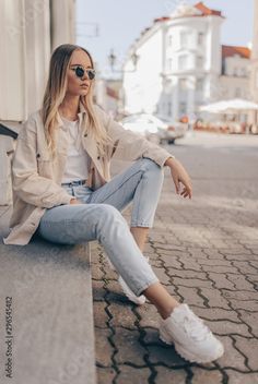 a woman sitting on the side of a building with her legs crossed and wearing white sneakers