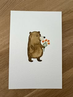 a brown bear holding flowers on top of a wooden table next to a piece of paper