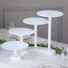 three tiered white cake stands on top of a table with greenery and wine glasses