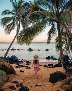 a woman in a pink dress and straw hat walks on the beach with palm trees