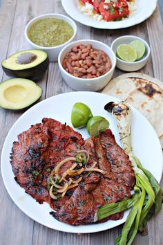 a white plate topped with meat next to bowls of beans and avocado on top of a wooden table