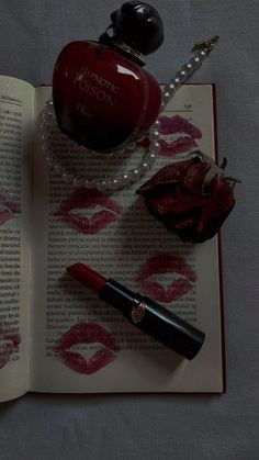 an open book with lipstick on it next to a bottle of perfume and pearl necklace