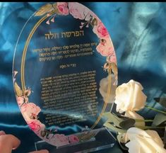 a glass plaque with pink flowers on it and writing in hebrew, surrounded by white roses