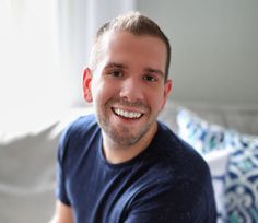 a smiling man sitting on a couch in front of a white wall with blue pillows