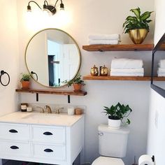 a white toilet sitting next to a bathroom sink under a round mirror and wooden shelves