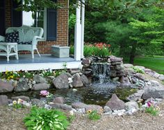 a small waterfall is in the middle of a rock garden area with chairs and flowers around it