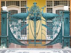 an iron gate with two peacocks on it in front of a yellow wall and windows