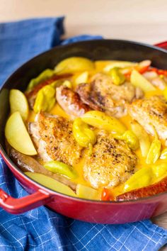 a red pan filled with chicken and vegetables on top of a wooden table next to a blue towel