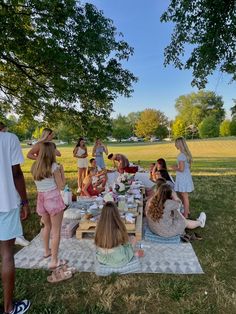 a group of people sitting at a picnic table in the park, eating and drinking