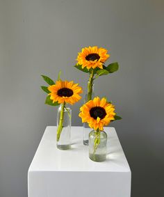 three vases with sunflowers in them sitting on a white table against a gray wall