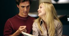 a young man and woman sitting next to each other looking at something on the table