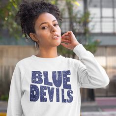 a woman in a blue devil sweatshirt is holding her hand up to her ear and looking off into the distance