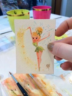 a hand holding up a card with a drawing of a girl on it and some paint brushes