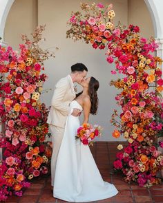 a bride and groom kissing in front of an archway with flowers on the wall behind them