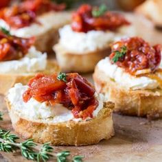 small pieces of bread topped with cheese and tomato sauce on a cutting board next to fresh herbs