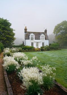 a white house sitting in the middle of a lush green field with lots of flowers