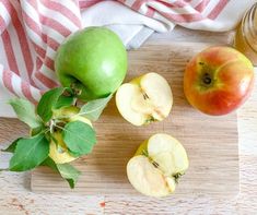 three apples on a cutting board next to a jar of honey and some green leaves