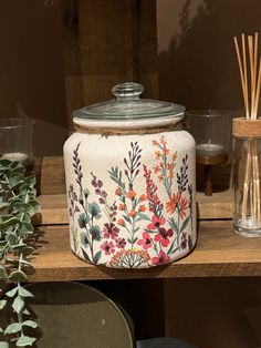 a jar with flowers painted on it sitting on a shelf next to candles and plants