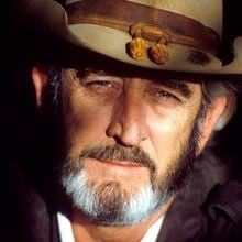 a close up of a person wearing a hat and suit with a beard in front of a black background