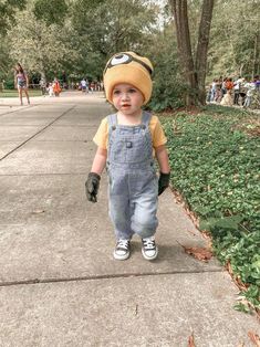 a young boy wearing overalls and a hat walking down a sidewalk with people in the background