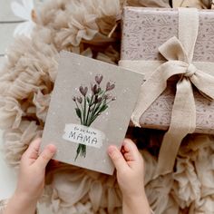 a person holding up a card with flowers on it next to two wrapped gift boxes