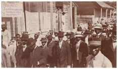 an old black and white photo of men in suits and hats walking down the street