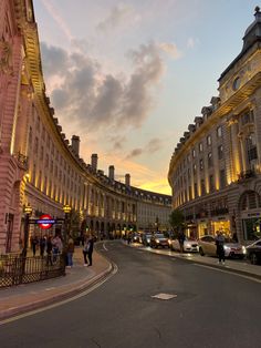 people are walking on the street in front of buildings at sunset or dawn with cars driving down the road