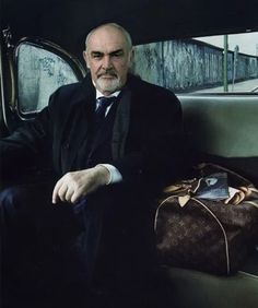 a man sitting in the back seat of a car next to a brown bag and purse