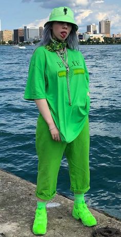 a woman in green is standing by the water with her hands on her hips and looking at the camera