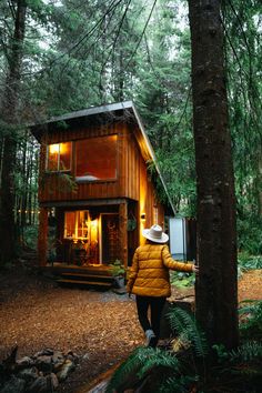 a person in a yellow jacket and hat walking towards a small cabin surrounded by trees