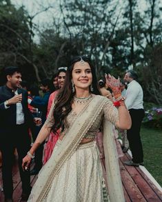 a woman in a white and gold lehenga is holding her hands out to the crowd