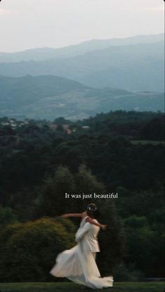 a woman in white dress flying through the air with trees and hills behind her on a cloudy day