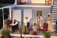 an outdoor stage with people dressed in period costumes and sitting on chairs, talking to each other