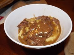 a white bowl filled with meat and gravy on top of a wooden table