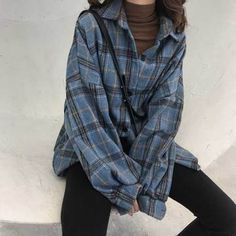 a woman sitting on the ground wearing black pants and a blue plaid shirt with her hands in her pockets