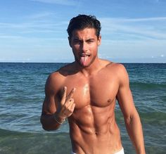 a shirtless man standing in the ocean with his tongue out and making a peace sign