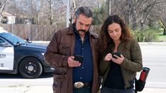 a man and woman standing next to each other while looking at their cell phones in front of a police car