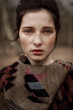 a young woman with freckled hair and blue eyes is wearing a sweater in the woods