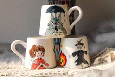 three coffee mugs with cartoon characters on them sitting next to each other in front of a lace doily