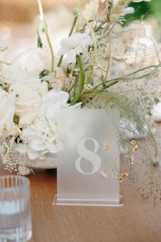 the table number is placed next to some white flowers and greenery in a vase