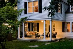 a white house with black shutters and chairs on the front porch at night time