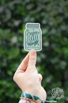 someone holding up a sticker with the words enjoy little things on it in front of some trees