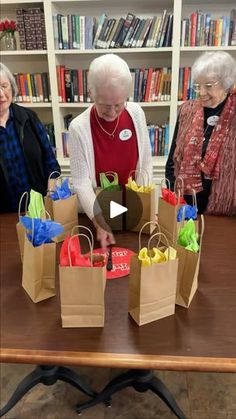 three older women standing around a table with bags on it and bookshelves in the background