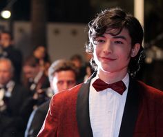 a young man wearing a red suit and bow tie at an event with people in the background