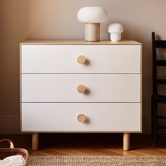 a white dresser with three drawers and two lamps on top, in front of a brown wall