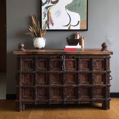 a painting hangs on the wall above a wooden cabinet with two vases and a potted plant