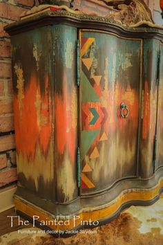 an old dresser painted with different colors and designs on it's sides, in front of a brick wall