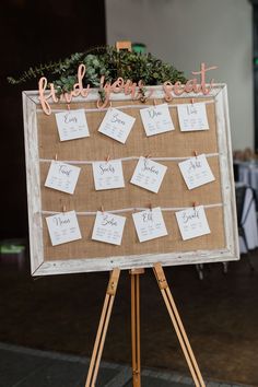 a wooden easer with seating cards attached to it and greenery in the center