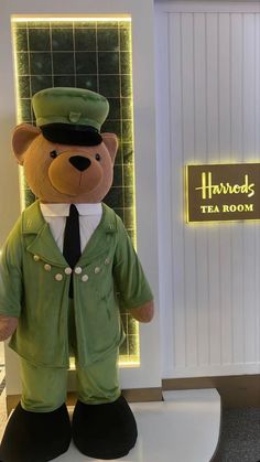 a teddy bear dressed in a green uniform standing on a white pedestal with a sign behind it that says harrison's tea room