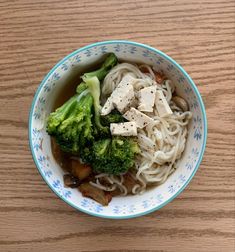 a bowl filled with noodles, broccoli and tofu on top of a wooden table